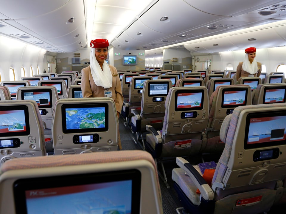 Zwei Flight-Attendants stehen im leeren Passagierraum, fotografiert von hinten, man sieht die Bildschirme jeweils an der Hinterlehne des Vordersitzes.