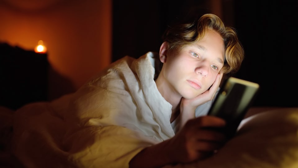 Eine junge Person liegt im Dunkeln und schaut aufs hell leuchtende Handy.