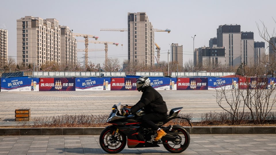 Ein Motorrad fährt von rechts nach links durchs Bild, im Hintergrund graue Hochhäuser und Baukräne.