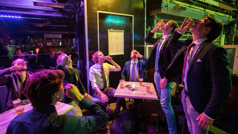 Eine Gruppe von sieben Männern trinkt vor einer Bar Shots mit Alkohol.