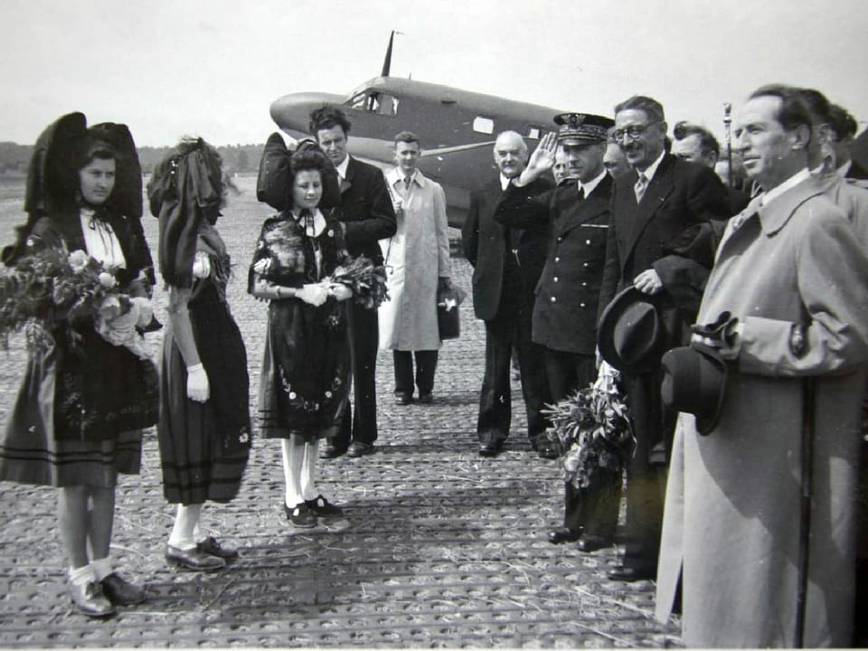 Auf einem historischen Bild stehen Politiker, Piloten und Frauen in Trachten vor einem Flugzeug.