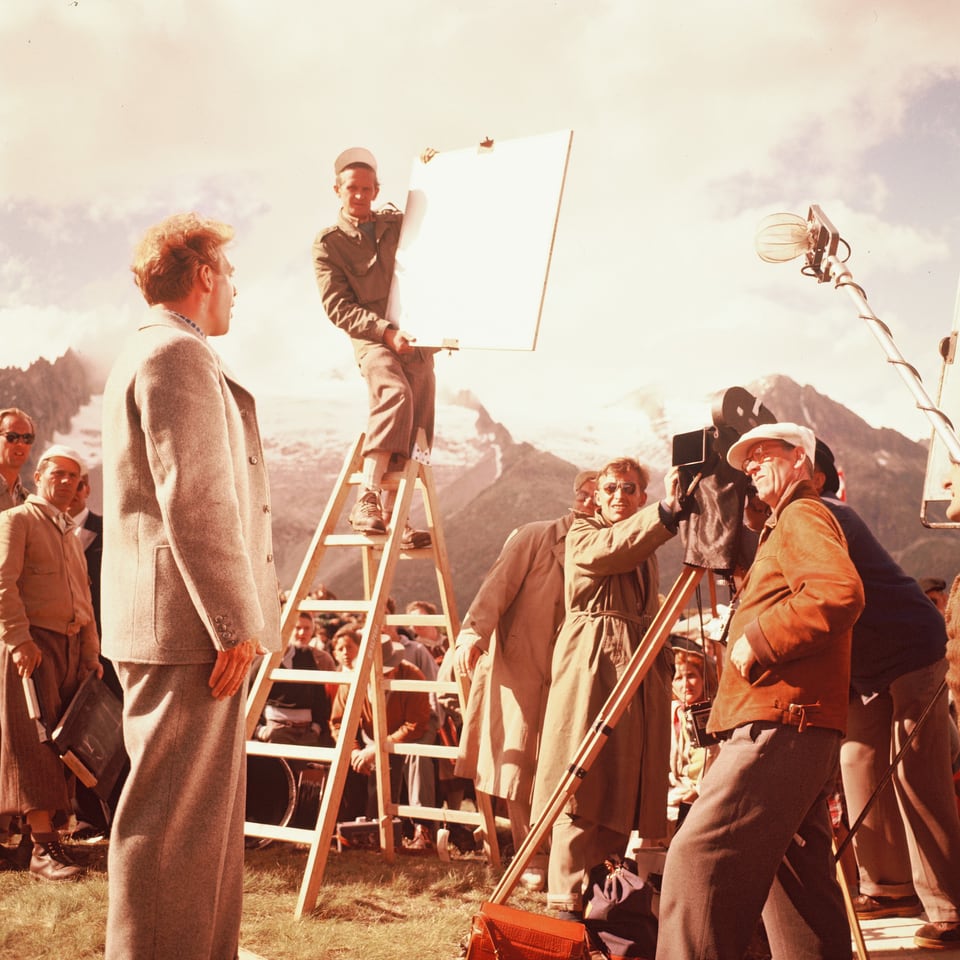 Hannes Schmidhauser steht gross vor der Kamera. Neben der Kamera Franz Schnyder, der Kameramann und der Kameraassistent. Auf einer Leiter neben der Kamera steht ein Beleuchhter mit einem Lichtreflektor. Hinter ihnen die Berge.