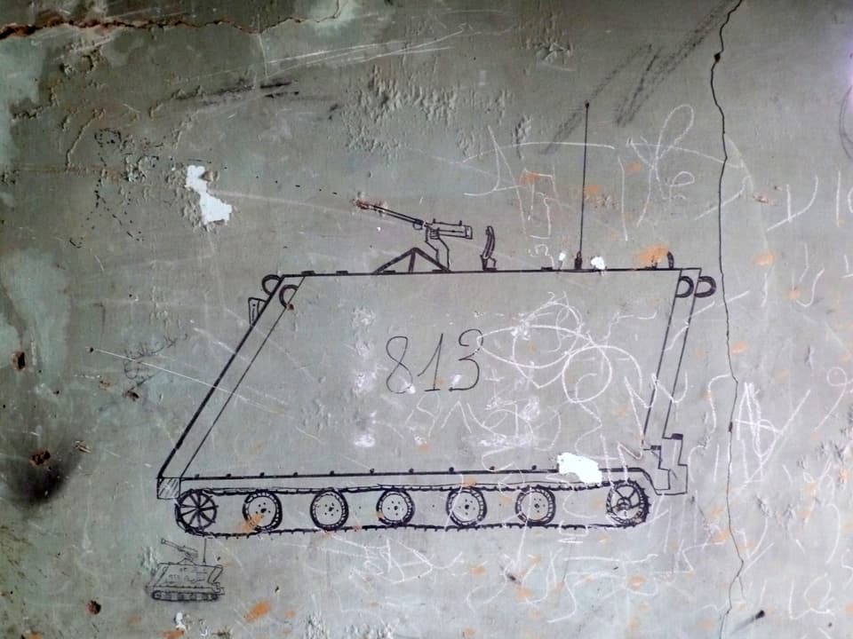 Eine Zeichnung eines schmalen Objektes auf Panzerrädern auf einer grauen, etwas verkritzelten Wand.