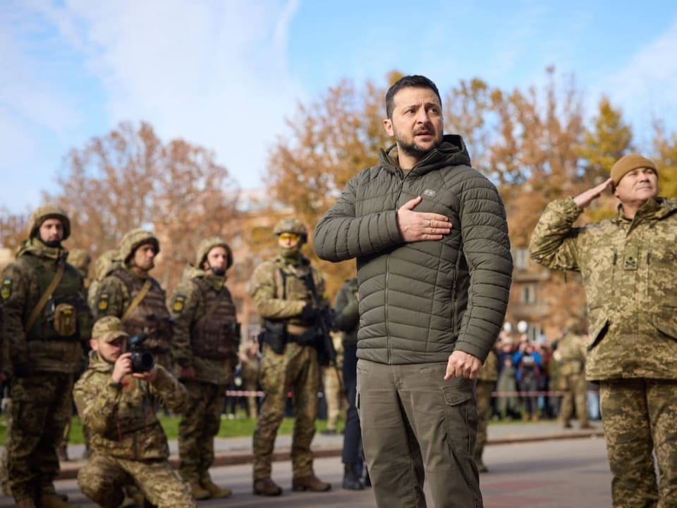 Selenski steht auf einem Platz und hält sich die rechte Hand auf die Brust. Hinter ihm stehen Soldaten.