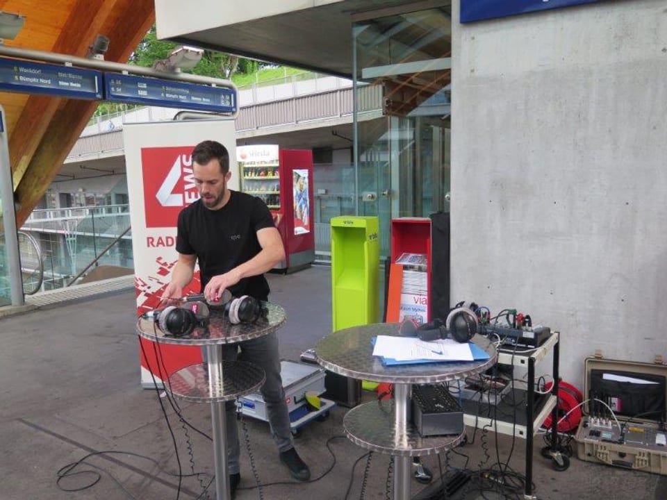 Ein junger Mann in schwarzem T-Shirt baut auf der Welle am Bahnhof Bern Mikrofone und Kopfhörer auf einem Stehtischchen auf. Im Hintergrund sind jede Menge Ausrüstungsteile zu sehen, sowie der Banner von SRF 4 News.