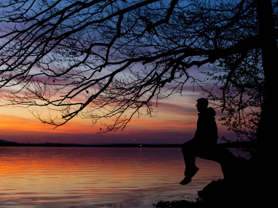 Oranger Himmel spiegelt sich in einem See. Ein Mann sitzt am Ufer unter einem Baum im Gegenlicht und geniesst die Stimmung