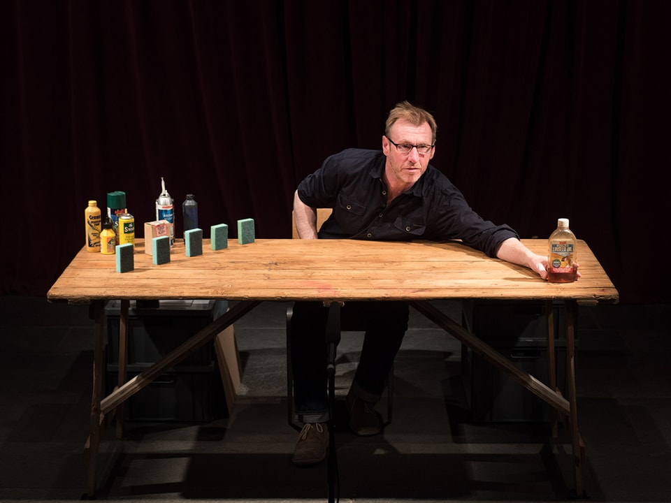 Theaterszene: Ein Mann sitzt an einem Holztisch und greift nach einer Ketchup-Flasche.