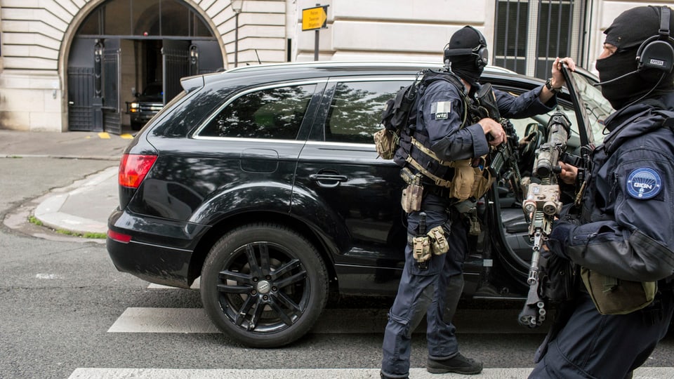 Zwei bewaffnete Männer vor einem schwarzen Auto.