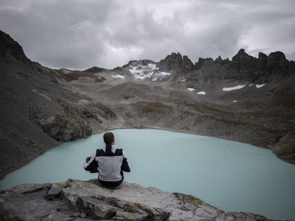 Person sitzt auf einem Felsvorsprung vor einem türkisfarbenen Bergsee und Bergen im Hintergrund.
