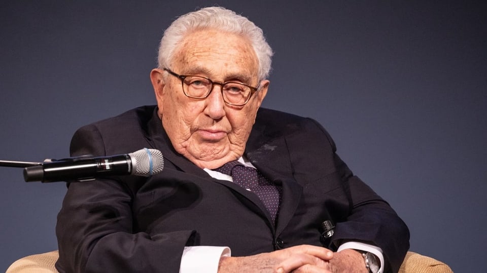 Henry Kissinger sitzt in einem Stuhl und hat ein Mikrofon vor dem Gesicht.