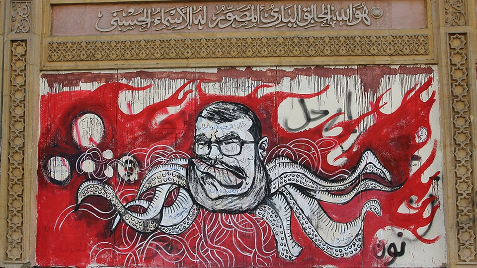 Abbildung eines Graffitos: Vor roten Flammen ist in schwarz-weiss der Kopf Mursis mit Brille abgebildet, von dem Tentakel-Arme abgehen.