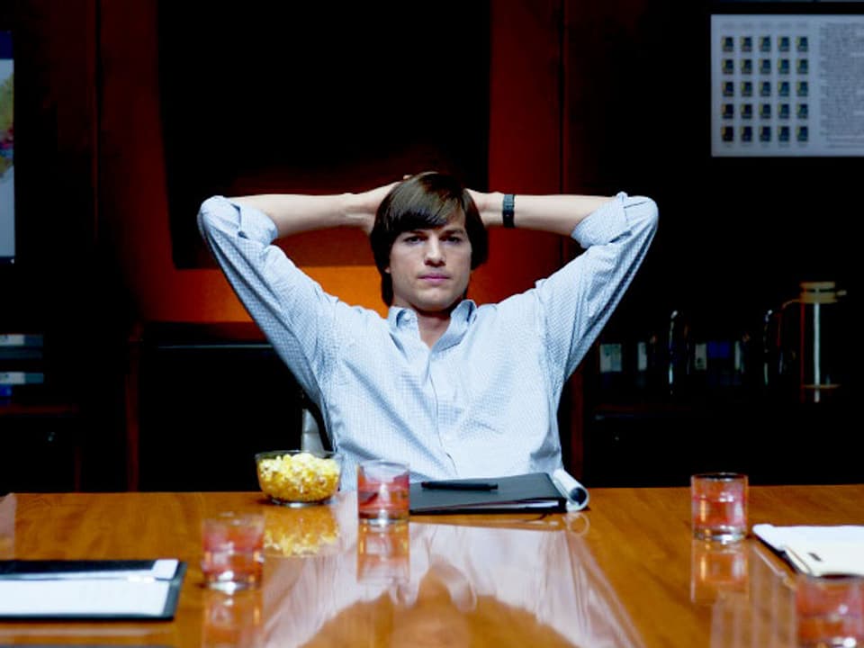 Asthon Kutcher sitzt als Steve Jobs mit hinter dem Kopf verschränkten Armen am Schreibtisch.