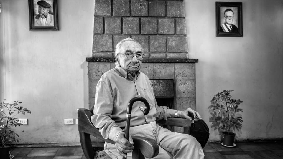 Alter Mann mit einem Gehstock in der Hand sitzt auf einem Sessel, hinter ihm Kamin, Zimmerpflanzen, Bilder an der Wand