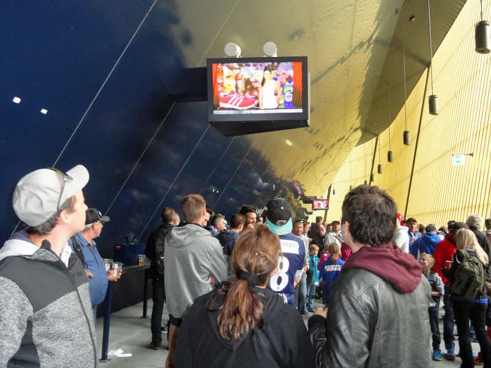 Personen stehen betrachten das Geschehen auf einem aufgehängten Bildschirm.