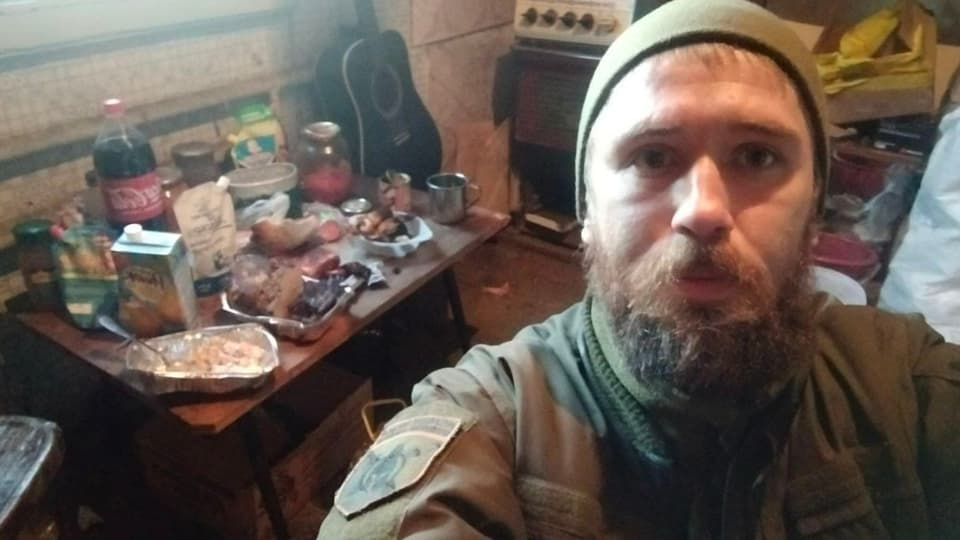 Mann in Militäruniform mit Mütze macht ein Selfie vor einem Tisch mit Essen und Getränken.