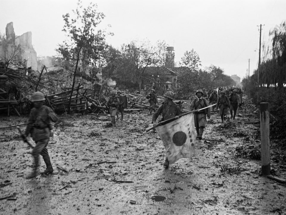 Japanische Soldaten gehen durch eine zerbombte Stadt.