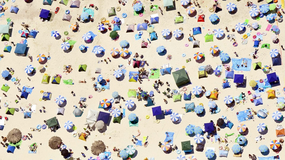 Flugaufnahme eines Sandstrands mit zahlreichen Sonnenschirmen, Zelten und Menschen auf Badetüchern.
