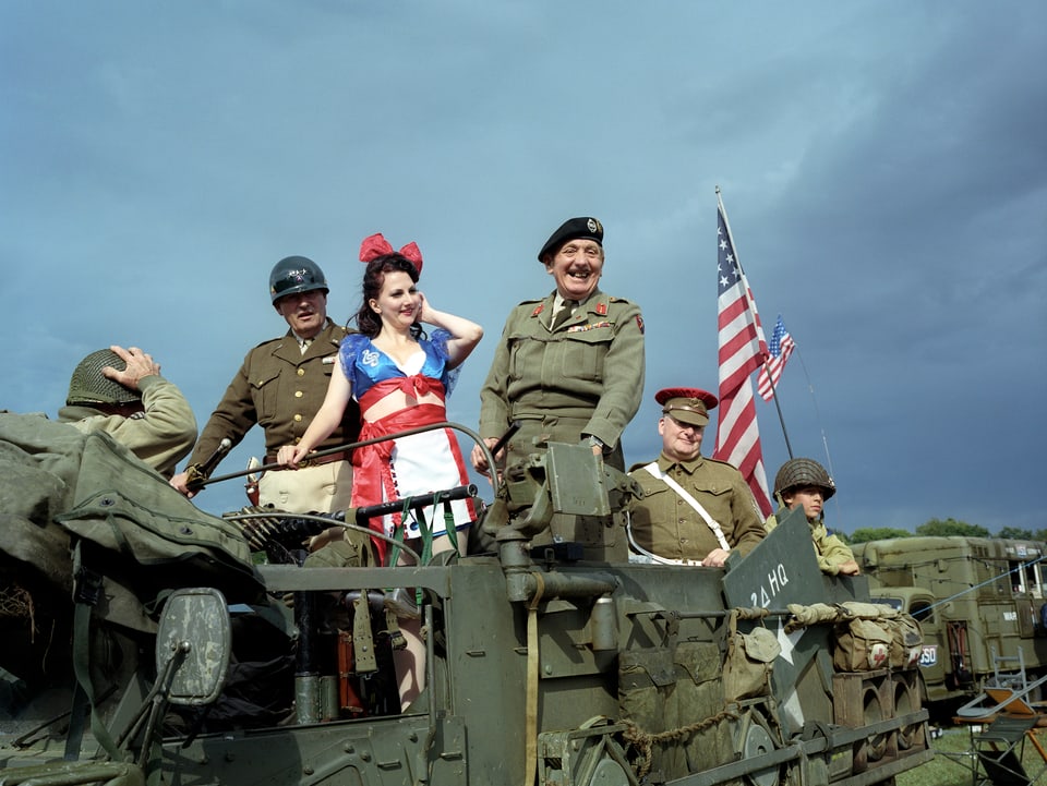 Soldaten stehen auf einem Panzer. In ihrer Mitte posiert eine Frau mit einem Kleid in den Farben der amerikanischen Flagge.
