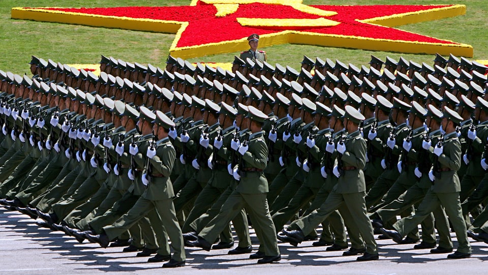 Im Gleichschritt marschieren hunderte chinesische Soldaten mit dem Gewehr im Anschlag. Im Hintergrund das Logo der Volksbefreiungsarmee.