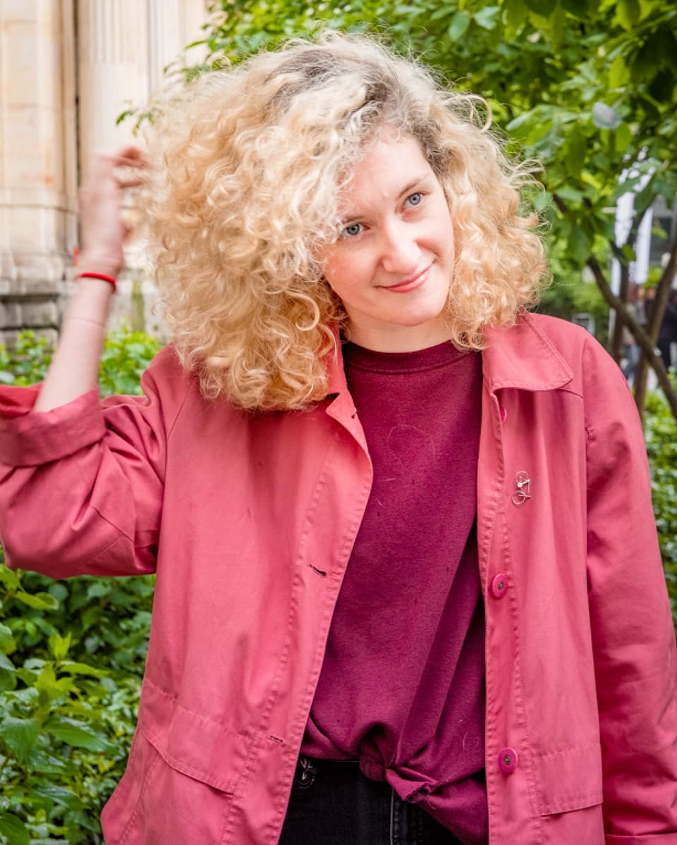 Eine junge Frau mit blonden, lockigen Haaren und rosa Jacke.