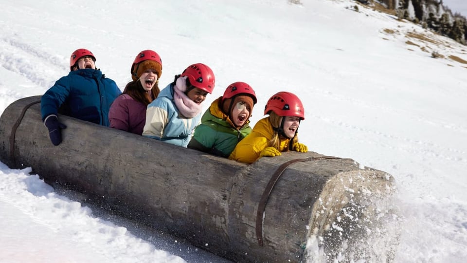 Fünf Kinder im Holz-Baumstamm fahren einen Schneehügel hinab.