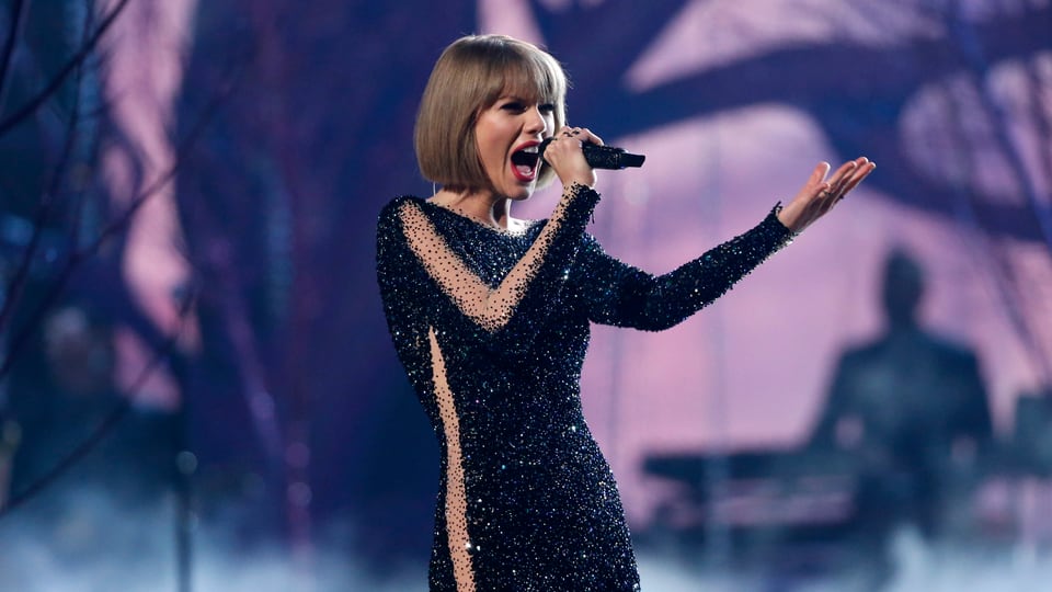 Taylor Swift singt in ein Mikrofon, im Hintergrund ein Bühnenbild mit Abbildung von Bäumen.