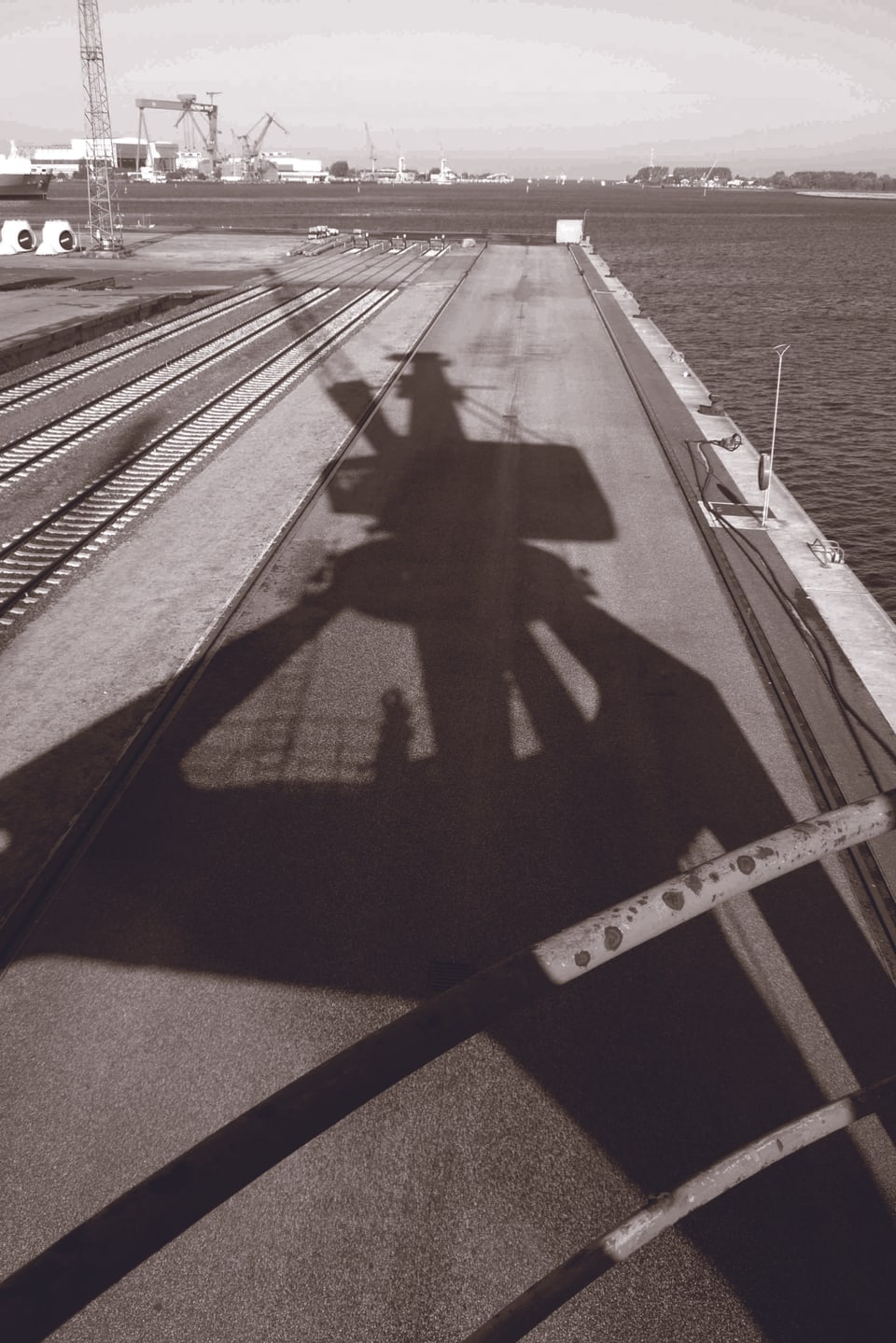 Der Schatten eines Hafenkrans in Schwarz-Weiss, dahinter das Meer.