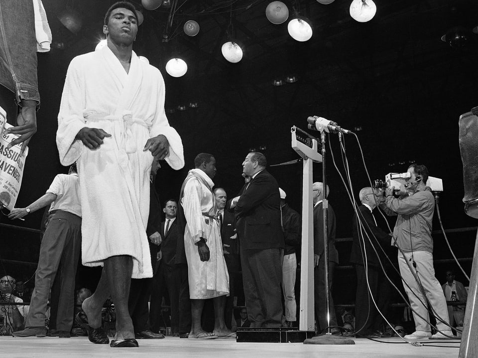 Ali im Vordergrund im weissen Bademantel vor dem Kampf gegen Sonny Liston am 25. Mai 1965, Liston im Hintergrund vor der Waage stehend