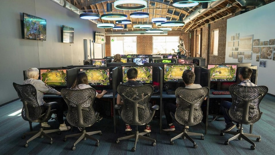 In einem Raum mit Tischen, Bildschirmen und Bürostühlen sitzen Jugendliche in einer Reihe und spielen ein Videogame