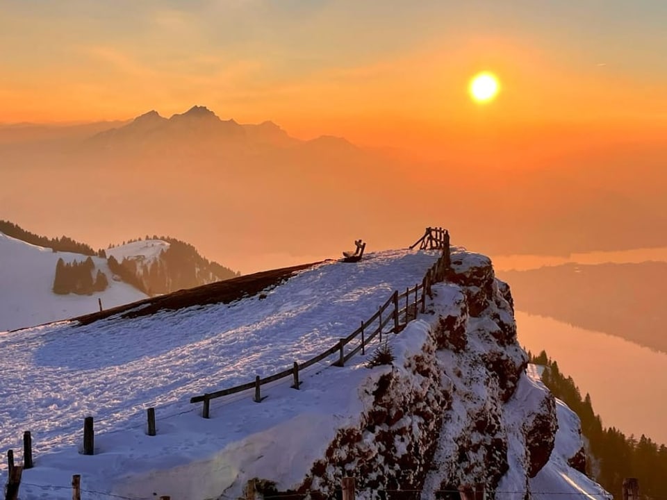 Blick von einem Berg mit Schnee ins Mittelland, wo die Sonne mit farbigem Himmel untergeht. 
