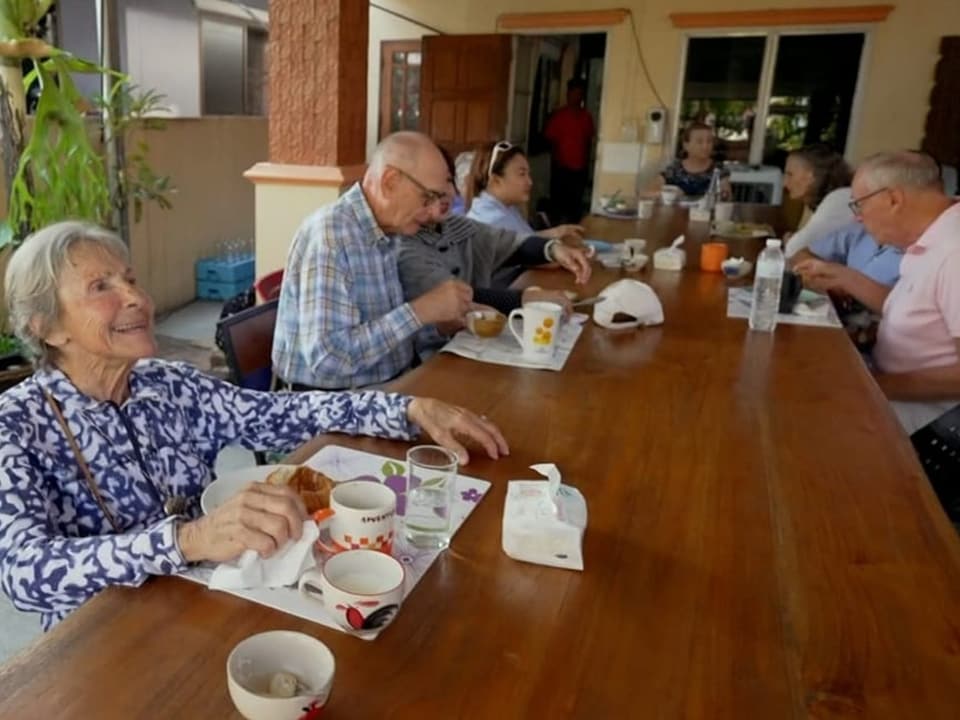 9 Personen sitzen an einem langen Tisch und essen Frühstück.