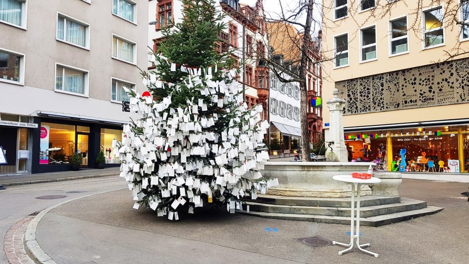 Tannenbaum auf Platz vor Brunnen, vollbehangen mit weissen Zetteln.