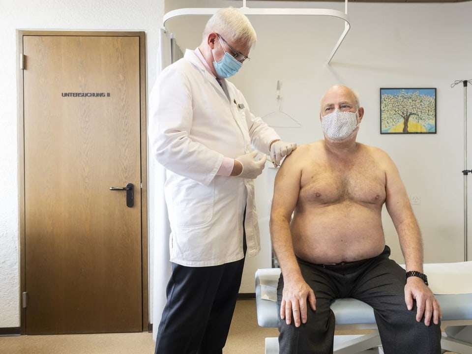 Ein Hausarzt impft einen älteren Mann mit nacktem Oberkörper. Beide tragen Masken.