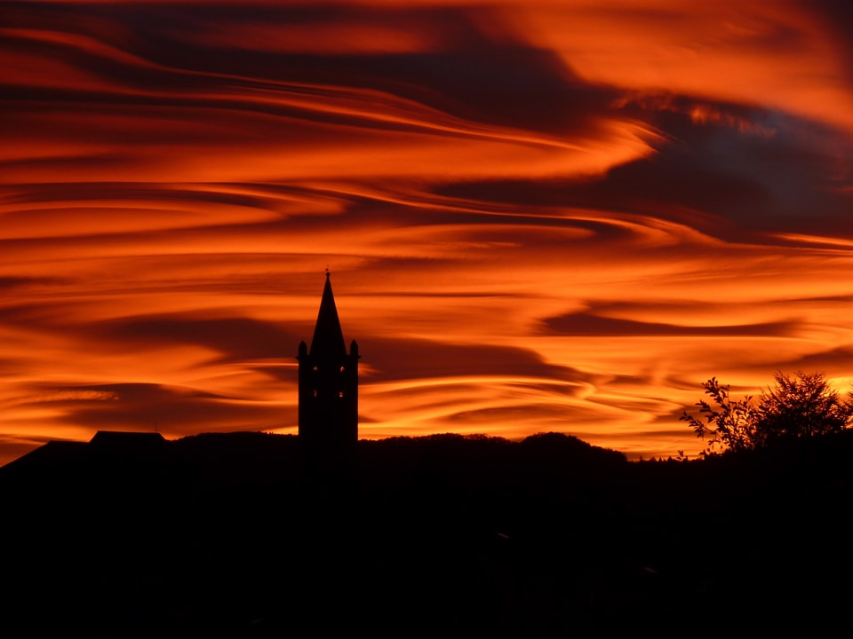 Im Vordergrund ein dunkler Kirchturm. Im Hintergrund sieht man den orangen bis roten Himmel mit spektakulären, ufoartigen Wolken.