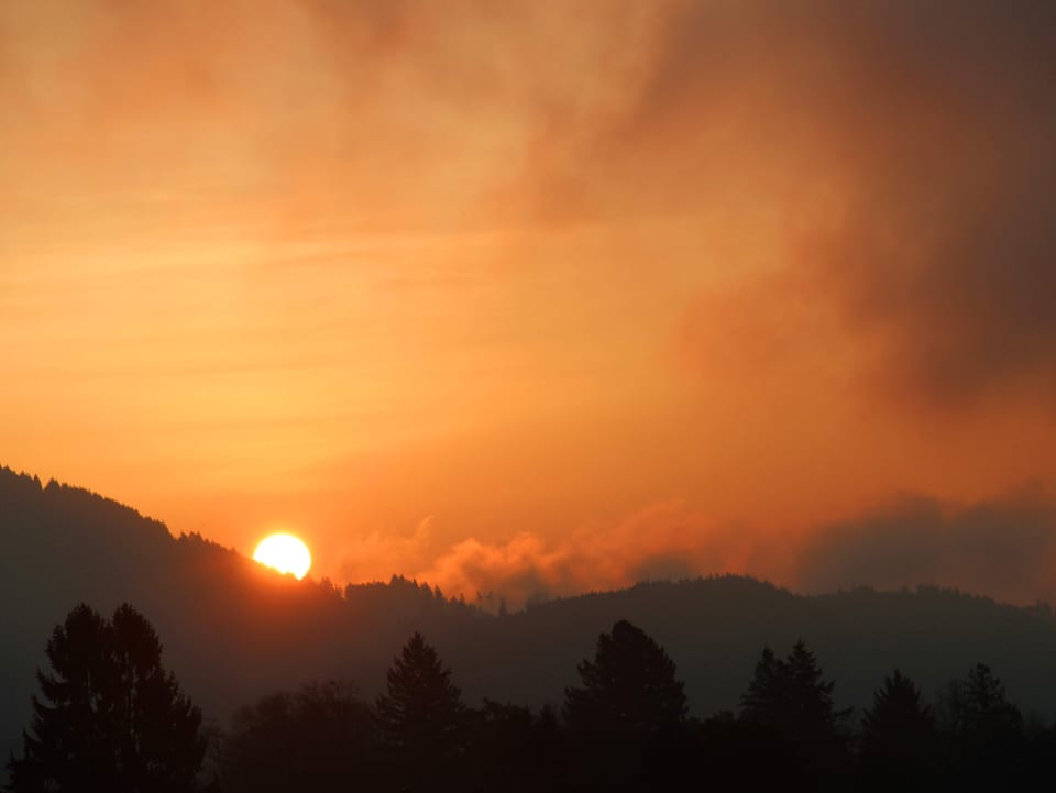 Die Sonne taucht hinter einem Hügel als roter Kugel auf. Im Tal hat es hochnebelartige Wolken.