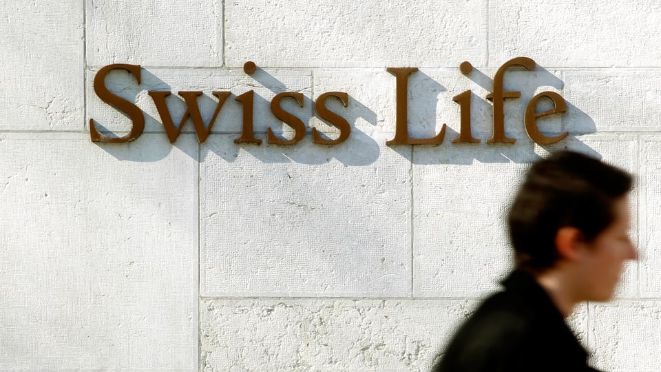 Emblem der Lebensversicherung «Swiss Life» an weisser Wand in Zürich. Eine Passantin schreitet daran vorüber.