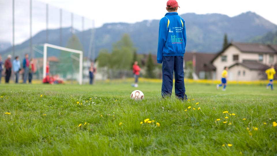 Ein Junge in einer blauen Jacke steht auf einem Fussballplatz, vor ihm liegt ein Fussball. Im Hintergrund steht ein Fussballtor, zudem hat es weitere Kinder.