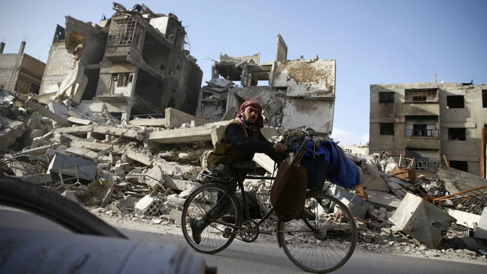 Einbeiniger Mann mit Turban fährt vor zerbombten Häusern mit dem Velo vorbei.