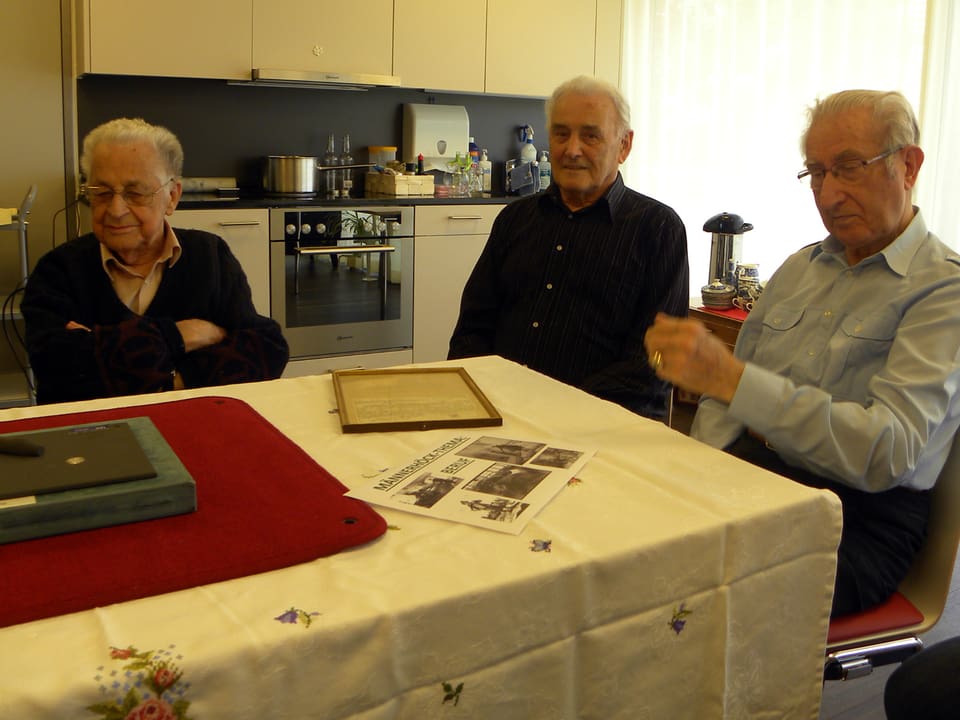 Drei alte Männer sitzen an einem Tisch in einer grossen Küche.