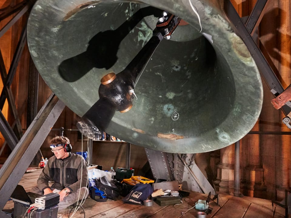Ein Mann mit Kopfhörern sitzt unter einer riesigen Glocke, die hin- und herschwingt.