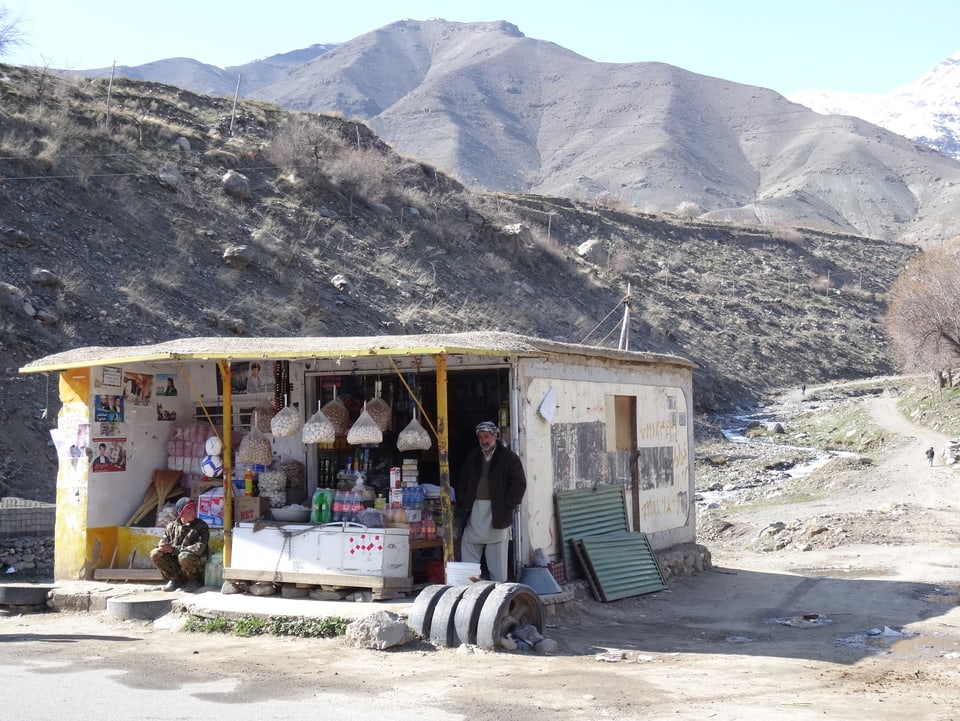 Ein kleiner Laden mit Auslegeware, davor zwei Männer; im Hintergrund karges Land.