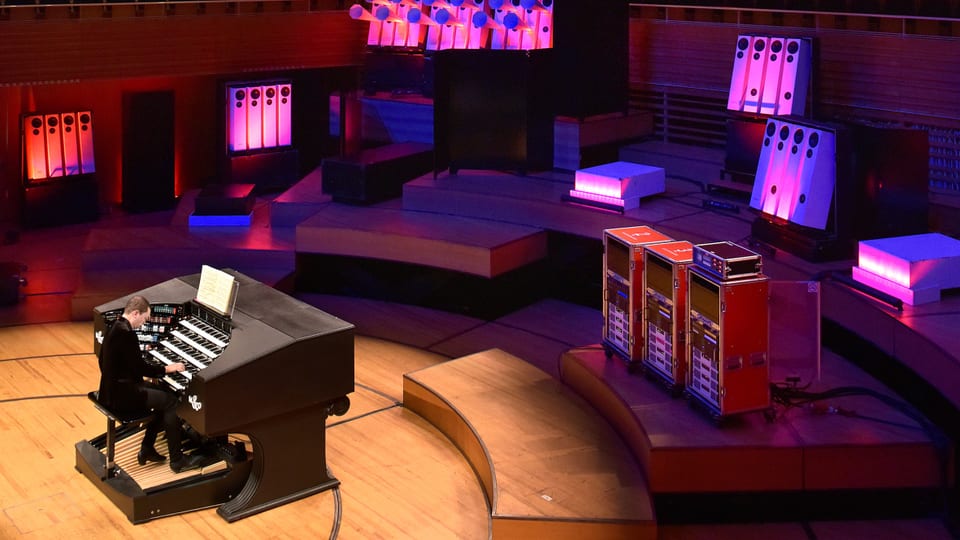 Ein Mann sitzt an einer Orgel, im Hintergrund grellpinke Lautsprecher.