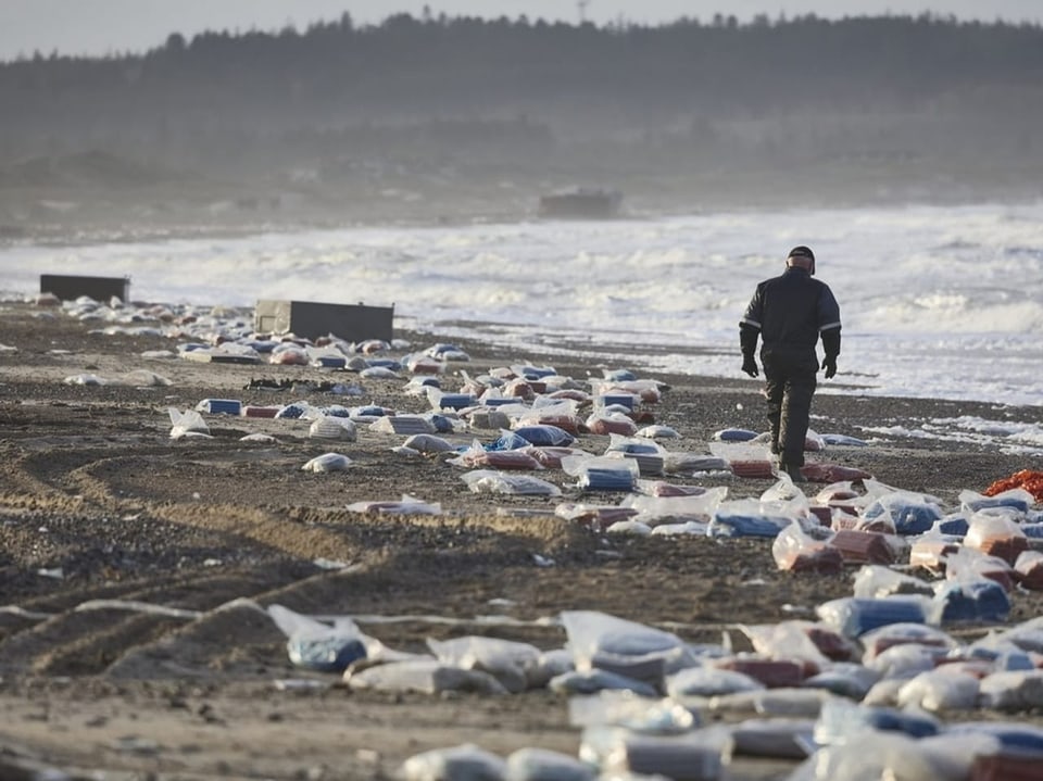Ein Mann läuft am Strand zwischen vielen gefüllten Plastiksäcken. Im Hintergrund liegen offenbar Kühlschränke.
