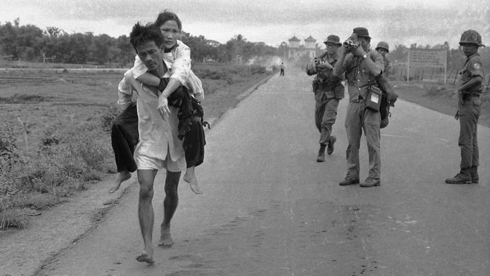Mann trägt Frau auf dem Rücken. Fotografen und Soldaten im Hintergrund.