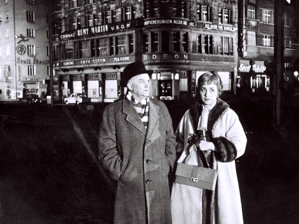 Ein älterer Mann und eine ältere Frau stehen Nachts auf dem Platz vor dem Café Odeon.