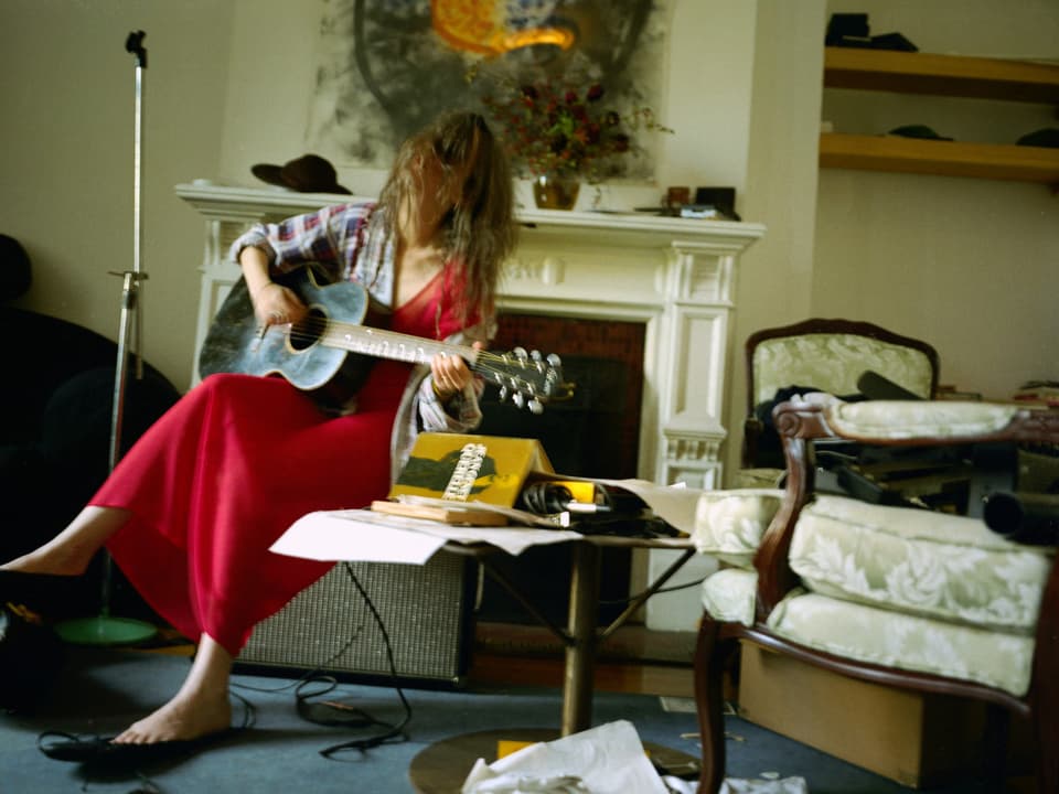 Patti Smith spielt in einem roten Kleid Gitarre, die Haare hängen ihr ins Gesicht.
