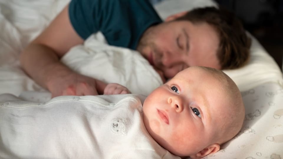 Ein waches Baby liegt neben einem schlafenden Vater im Bett