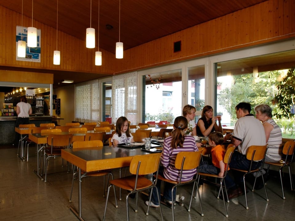 Menschen sitzend und sprechend in einer Cafeteria mit Holzverkleidung und hängenden Lampen.