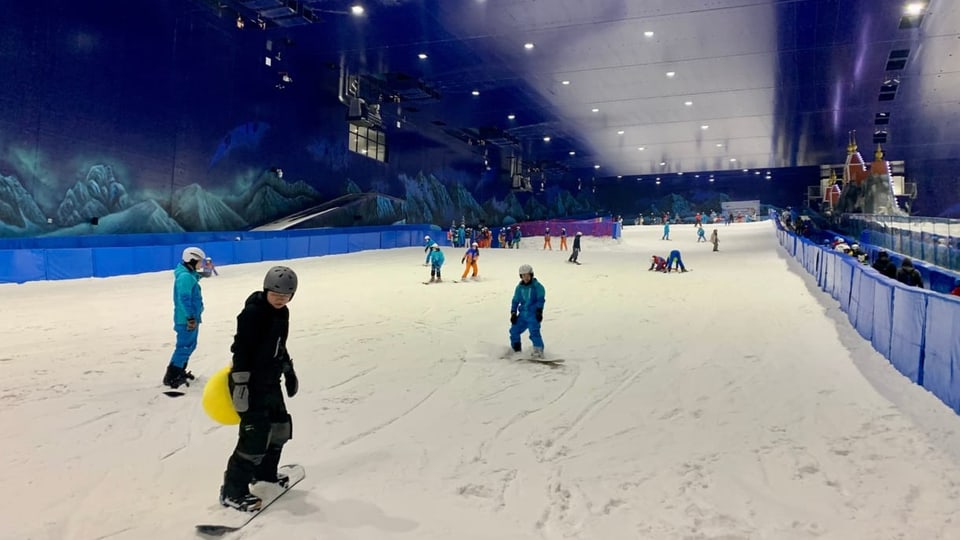 In einer Indoor-Halle fahren Menschen auf Snowboards und Skiern den Schneehang hinunter.