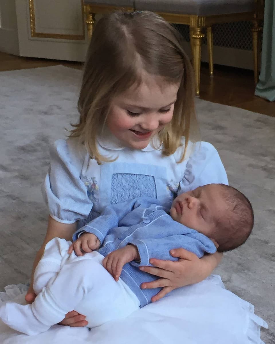 Prinzessin Estelle sitz in einem weiss-blauen Kleidchen am Boden und hält ihren Baby-bruder Oscar im Arm. 